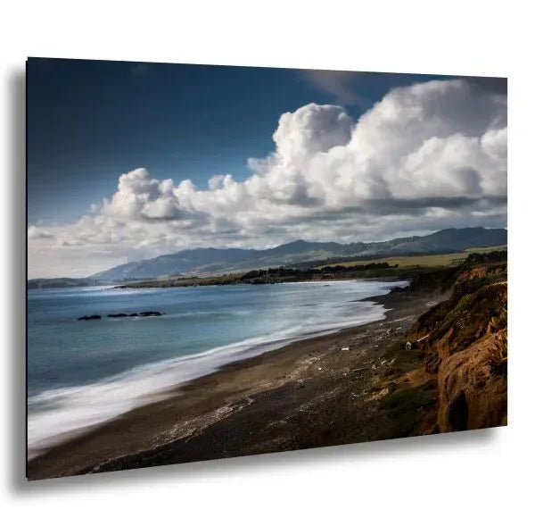 Coastal landscape with waves gently washing onto the shore, surrounded by green hills and dramatic clouds above the ocean.