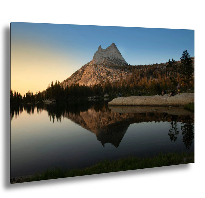Mountain peak reflected in a calm lake at sunset, with trees surrounding the water and a group of people relaxing on the rocky shore.