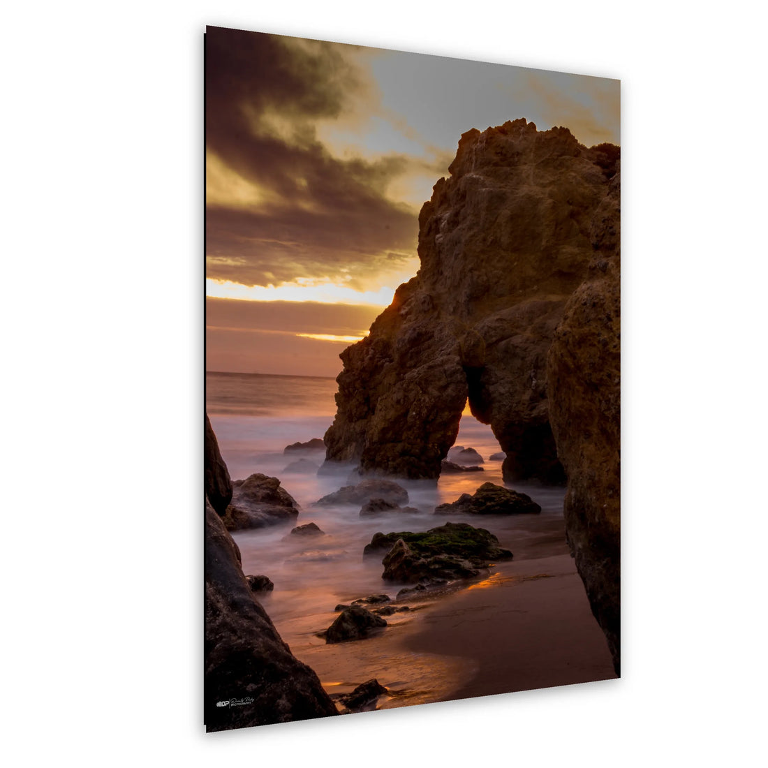 Golden sunset over a rocky coastline with waves crashing against the shore, highlighting an arch-shaped rock formation and soft clouds above.