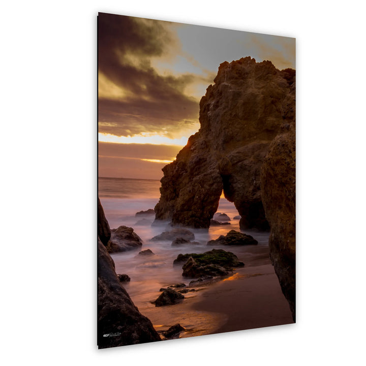 Golden sunset over a rocky coastline with waves crashing against the shore, highlighting an arch-shaped rock formation and soft clouds above.