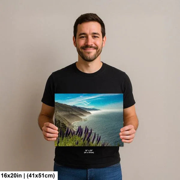 Man holding a 16x20 inch print of a scenic coastal view with purple flowers in the foreground, overlooking the ocean and distant mountains under a clear blue sky.