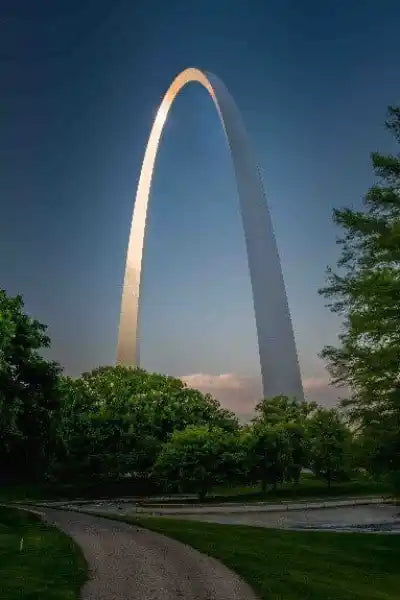St. Louis Gateway Arch at dusk with lush green trees and dramatic blue sky.