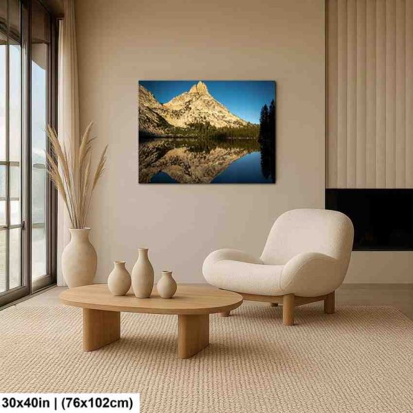 Mountain lake reflection landscape canvas above cream chair in minimalist modern living room with wooden furniture.
