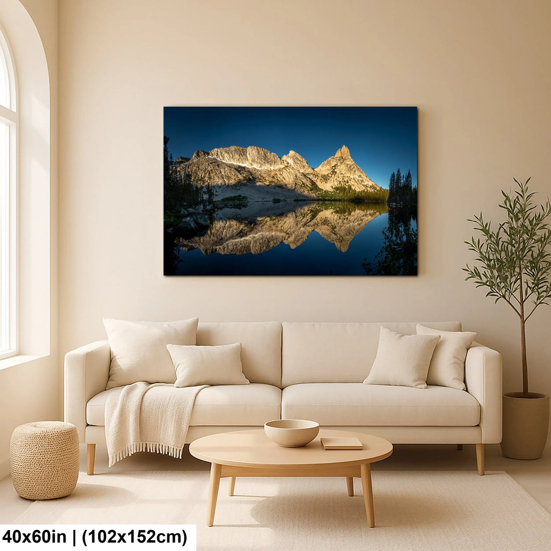 Large canvas print of a mountain range and its reflection in a serene lake, displayed above a cozy sofa in a minimalist living room with natural light.