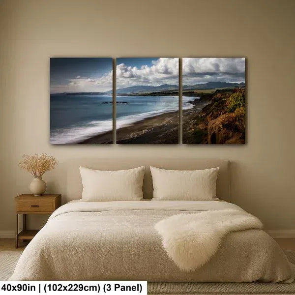 Coastal landscape triptych artwork above a cozy bed, featuring waves, sandy beach, and lush hills under dramatic skies in a calming bedroom setting.