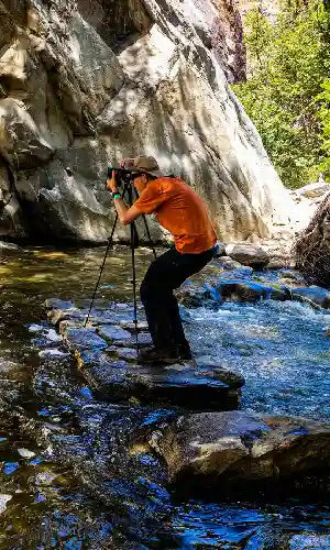 Photographer on rocks capturing waterfall in forest stream with camera and tripod.